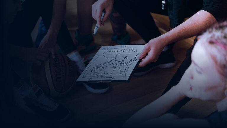 Trainer skizziert auf einem Klemmbrett Basketball-Spielzüge, umgeben von jungen Spielerinnen und Spielern in der Auszeit.