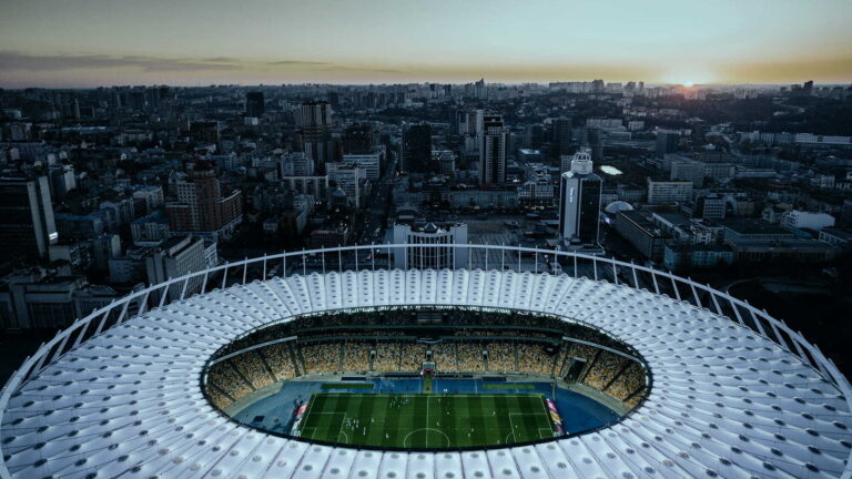 Luftaufnahme eines großen Rundstadions mit Fußballfeld bei Sonnenuntergang, im Hintergrund die Skyline der Stadt.