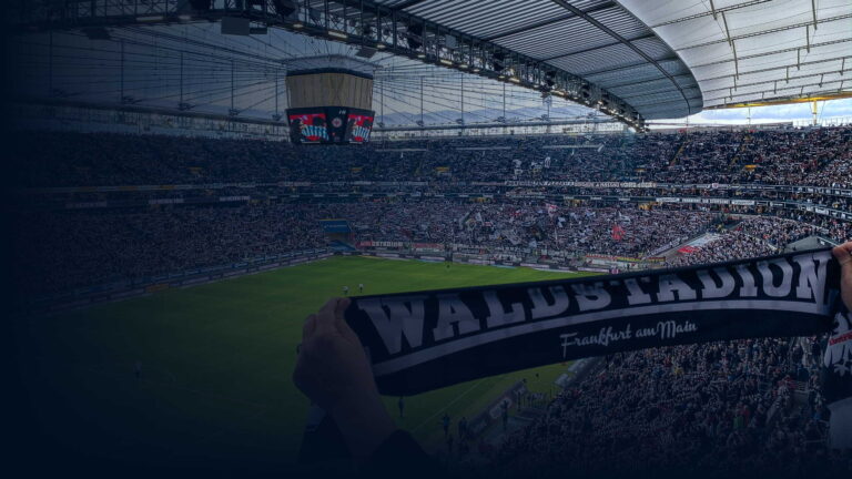 Blick von der Tribüne ins vollbesetzte Frankfurter Waldstadion, im Vordergrund ein Fanschal mit der Aufschrift Waldstadion Frankfurt am Main.